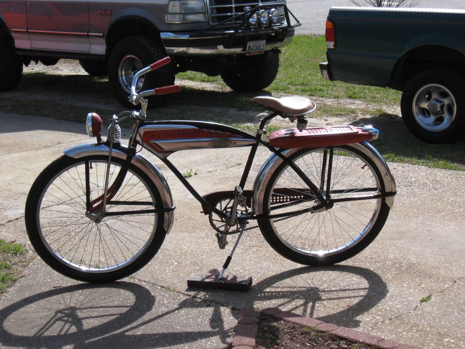 Vintage bicycle with red and chrome frame, tan seat, and red rear rack, parked on concrete. Two trucks are visible in the background