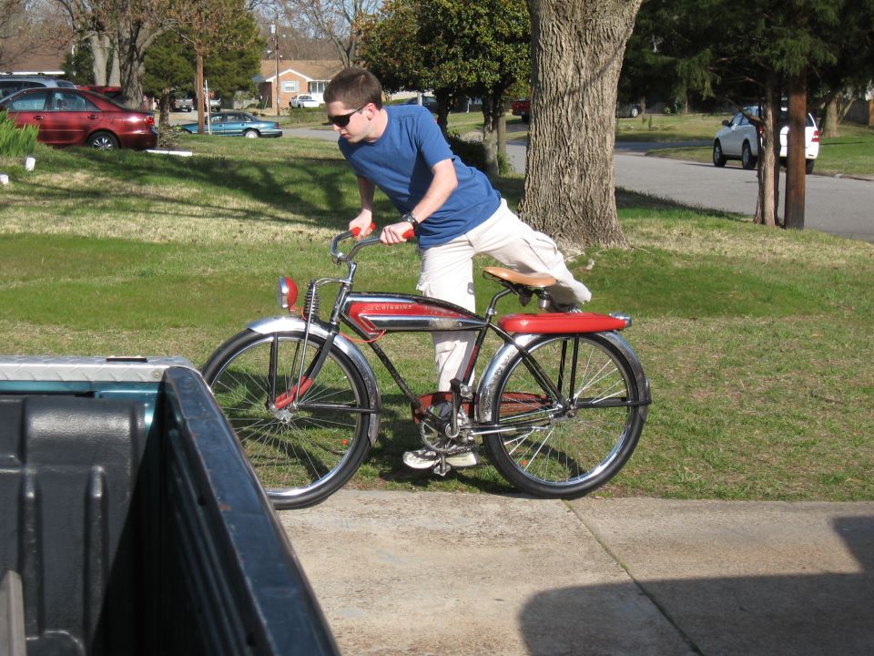 Man in blue shirt and khaki shorts pushing a red and chrome vintage bicycle on a sidewalk