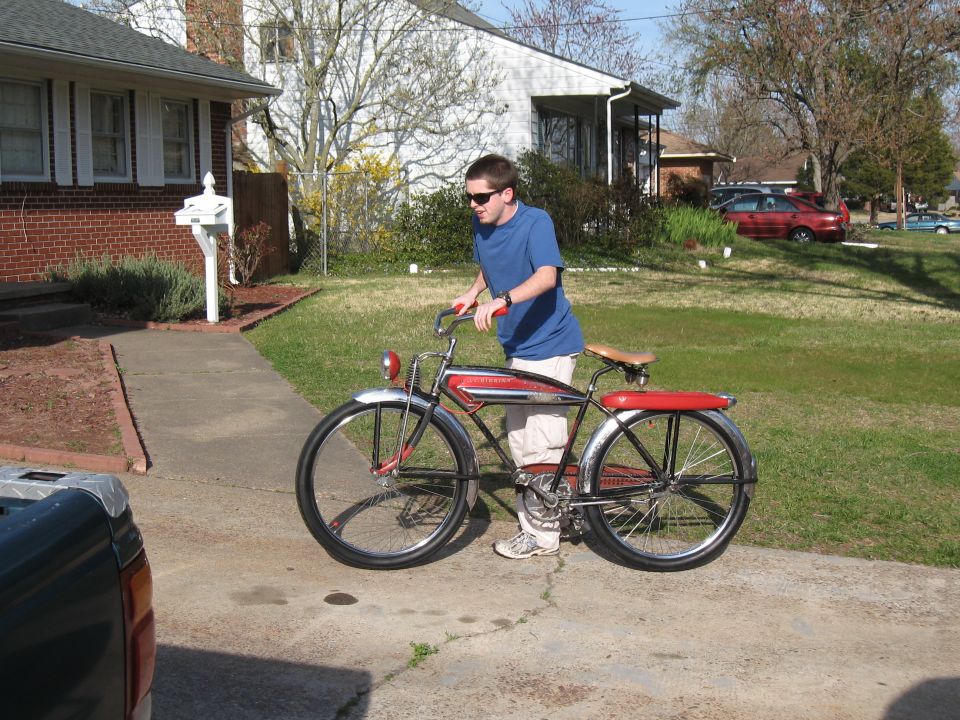 guy with sunglasses in blue shirt and tan pants standing next to a red and black vintage bicycle with chrome fenders and a headlight