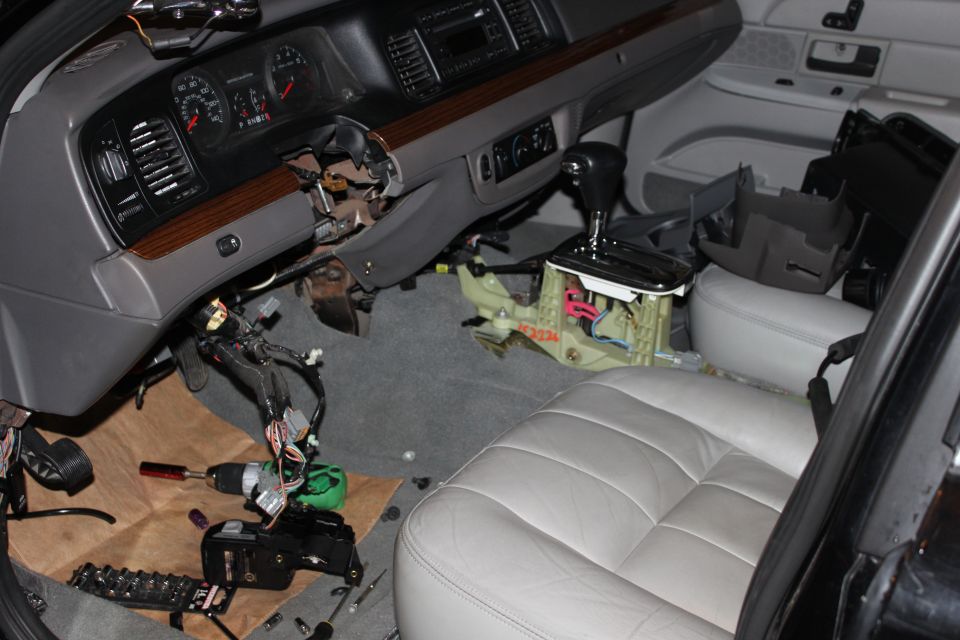 Car interior with dashboard and center console disassembled, showing wiring and parts. Tools are on the floor