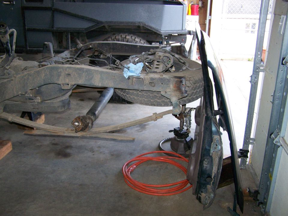 Vehicle frame on jack stands, drive shaft removed, door leaning against it. Orange hose on floor