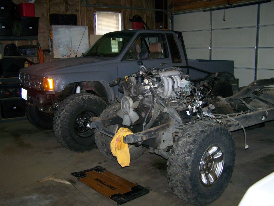 Gray pickup truck in garage, engine removed and chassis exposed. Another truck is parked behind it