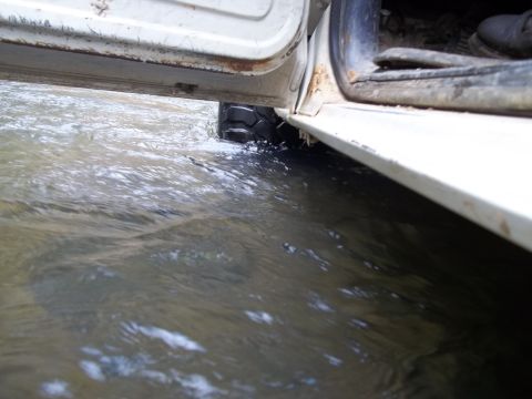 1990 Ford Bronco - A white vehicle drives through shallow water with the door open.