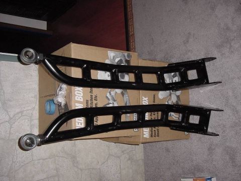 1987 Ford Bronco - Two black control arms with visible bearings are placed on a cardboard box, with a SuperMotors logo at the bottom right.