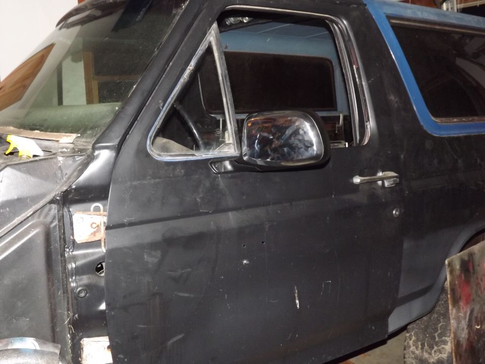 Black Ford Bronco door and fender, showing chrome mirror and door handle. The vehicle appears to be undergoing restoration