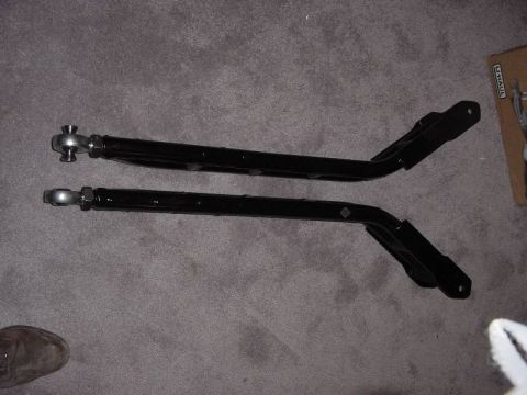1987 Ford Bronco - Two black control arms with spherical joints are laid out on a carpeted surface.