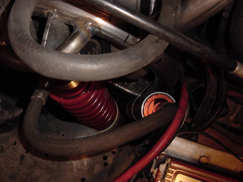 Red coilover shock with gold threaded collar amid hoses and steel tubing in an engine bay