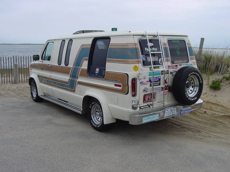 Cream 1980s Ford conversion van, rear 3/4 view, with stripes, spare tire and stickers at a beach.