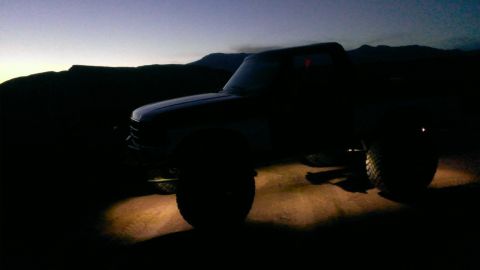 1994 Ford Bronco - A lifted pickup truck is parked on a dirt road as the sun sets, with its headlights casting a glow on the ground.