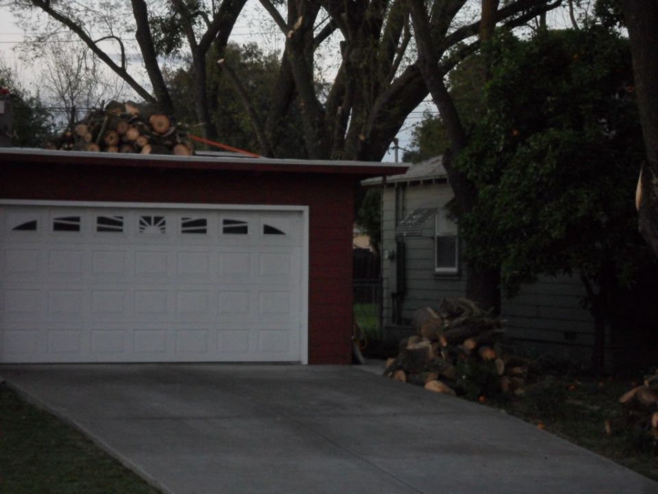 Garage with white door and red trim, driveway, cut logs on roof and beside house