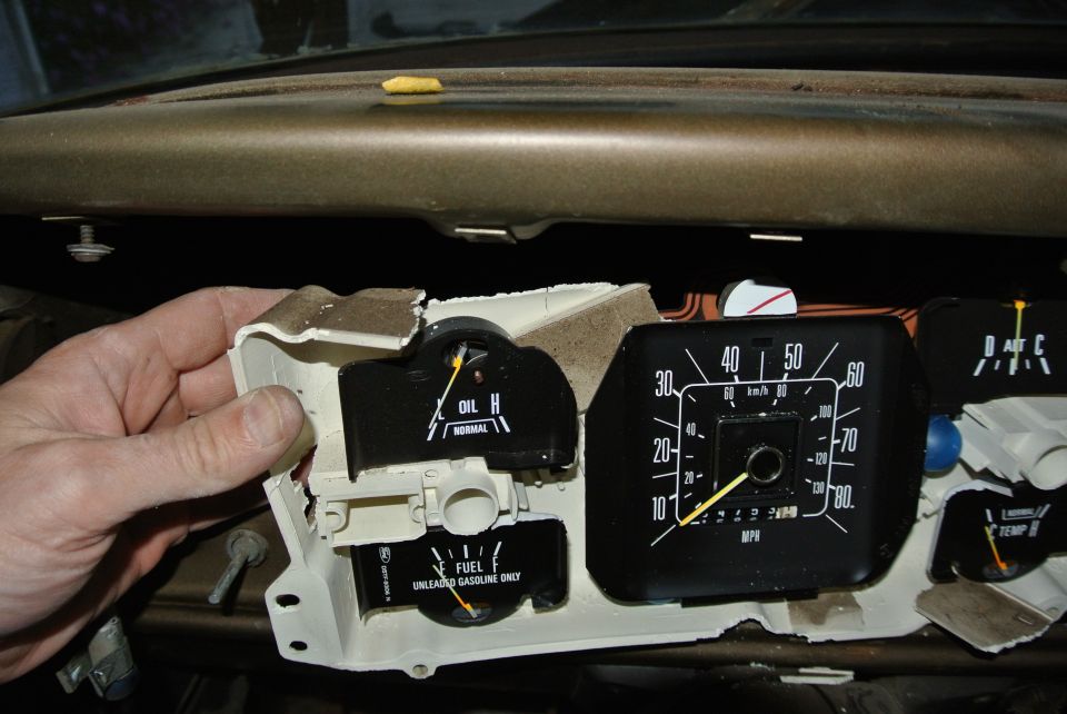 Hand holding a car's instrument cluster with oil and fuel gauges, plus a speedometer