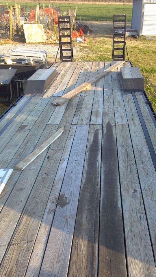 Empty wooden trailer deck with two metal ramps at the back. A wooden plank lies across the deck