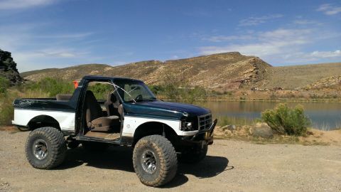 1994 Ford Bronco - Modified Ford Bronco sits parked beside a calm lake under a bright blue sky with distant hills.