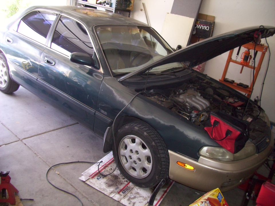 Dark green sedan with hood open, tan lower front bumper, and a red tool bag on the fender