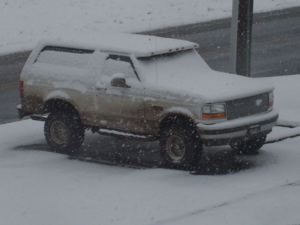 Snow-covered white lifted Ford Bronco parked roadside during a snowstorm