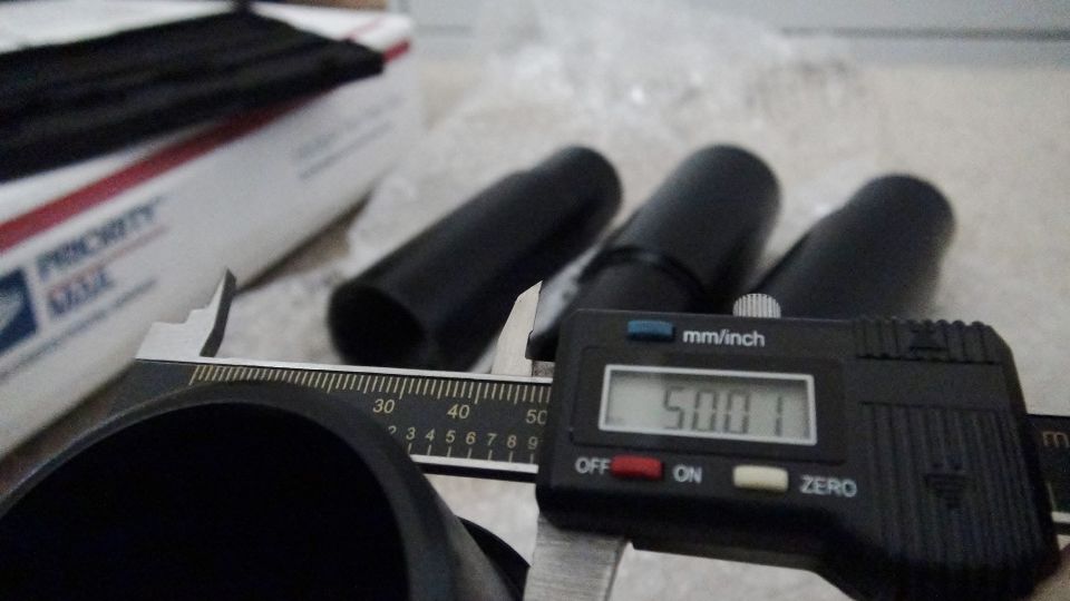 Digital caliper measuring 50.01mm on black cylindrical parts, with Priority Mail box in background
