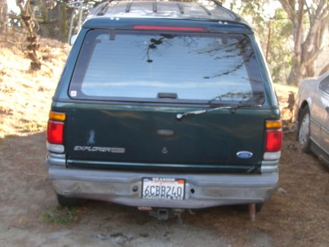 1973 Ford Ranchero - Rear view of a dark green Ford Explorer Sport Trac with a camper shell, parked outdoors next to another vehicle.