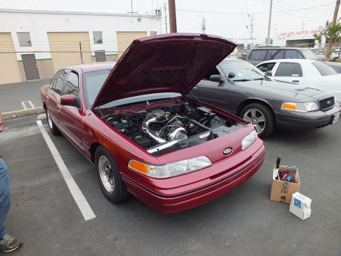 2003 Ford Crown Victoria - Red first-gen Ford Crown Victoria with raised hood and supercharged engine parked beside a gray Crown Victoria.