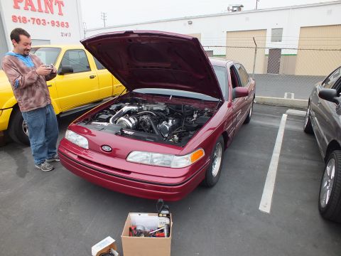 2003 Ford Crown Victoria - Person holding a camera next to a red Ford Crown Victoria with raised hood; a yellow Ford truck and paint-shop sign behind.