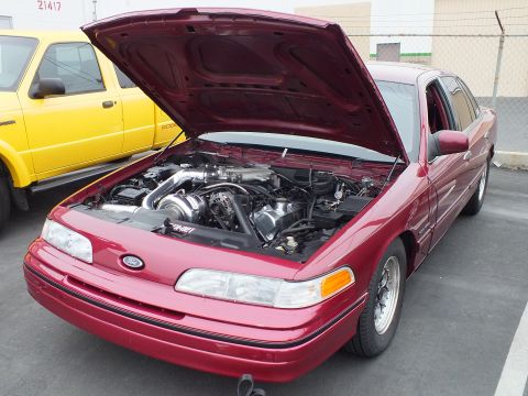 2003 Ford Crown Victoria - Red Ford Crown Victoria sedan with open hood revealing a belt-driven supercharger, beside a yellow truck.