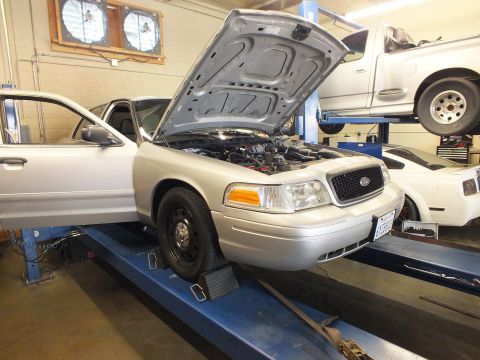 2003 Ford Crown Victoria - Silver Crown Victoria with steel wheels and open hood sits on a shop alignment rack, tire on a turn plate.