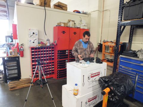 2003 Ford Crown Victoria - Technician handles components atop Hughes boxes beside red parts cabinets, a tripod and a fire extinguisher.