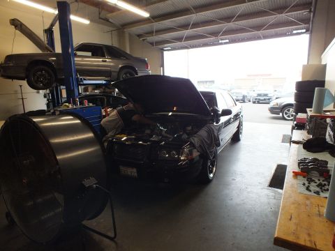 2003 Ford Crown Victoria - Black Crown Victoria being serviced with hood open in a shop bay; a silver SUV sits on a lift behind a large fan.