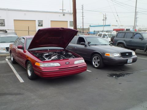 2003 Ford Crown Victoria - Red first-gen Crown Victoria with open hood parked next to a gray later-gen Crown Victoria outside a shop.