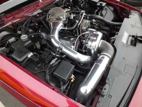 2003 Ford Crown Victoria - Bright polished intake piping routes to a centrifugal supercharger in a red vehicle engine bay