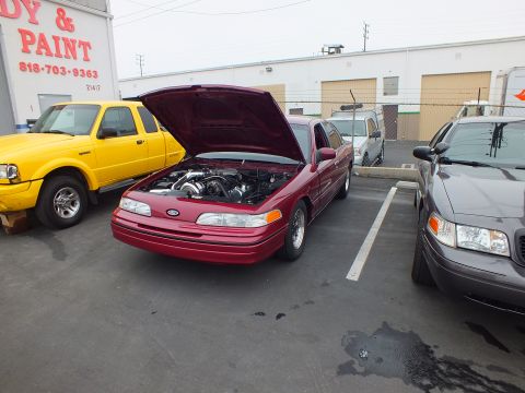 2003 Ford Crown Victoria - Maroon Ford sedan parked hood-up outside an auto body and paint shop, flanked by a yellow Ranger pickup