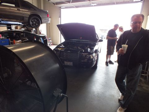 2003 Ford Crown Victoria - Black sedan with raised hood in a garage, a big drum fan at left and people standing near the door