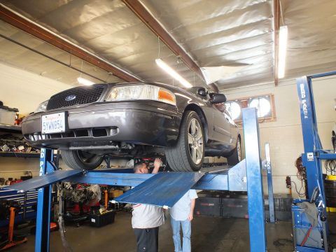 2003 Ford Crown Victoria - Crown Victoria up on a four-post lift, two techs working underneath, Ford oval visible on grille