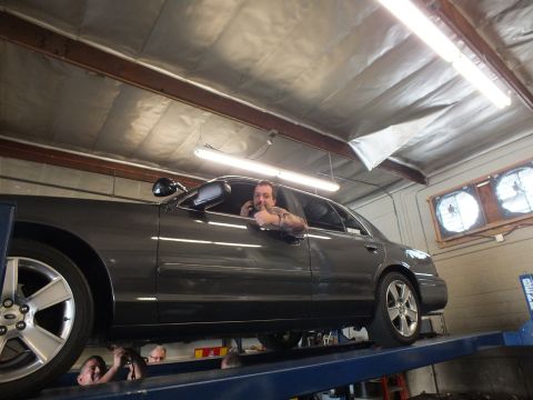 2003 Ford Crown Victoria - Man sits in a raised gray sedan on a lift, arm out the window holding a phone, in a garage bay