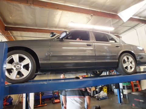 2003 Ford Crown Victoria - Side profile of a gray sedan elevated on a shop lift with mechanics working at floor level