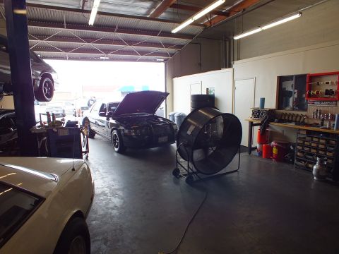 2003 Ford Crown Victoria - Repair shop interior with a black sedan hood-up at the door, a big drum fan and parts wall