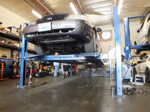 2003 Ford Crown Victoria - Mechanic works beneath a gray Ford sedan raised on a blue BendPak lift in a busy garage bay
