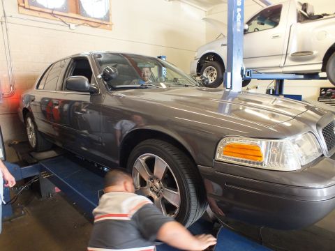2003 Ford Crown Victoria - Driver sits in a gray Ford sedan on an alignment rack as a worker crouches near the front fender