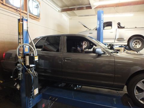 2003 Ford Crown Victoria - Gray full-size sedan sits on a shop rack while a person inside checks the cabin, lifted truck in back