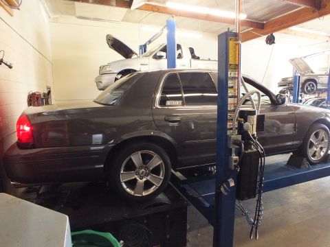 2003 Ford Crown Victoria - Rear quarter of a gray Ford sedan raised on a two-post lift with hood open in a repair shop