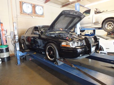 2003 Ford Crown Victoria - Black full-size sedan with open hood and front push bar sits on a blue alignment rack indoors