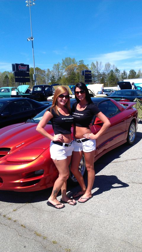 1980 Pontiac Trans Am - Two women stand next to a red sports car in a parking lot with other vehicles visible.