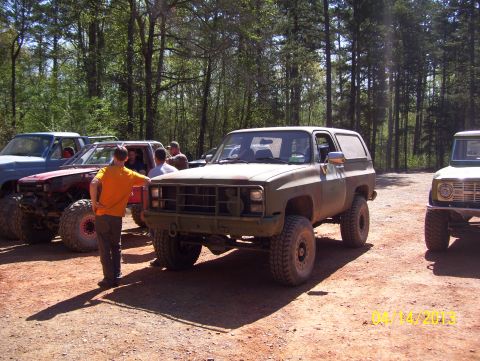1990 Ford Bronco - Several off-road vehicles are parked on a dirt lot surrounded by trees, with people gathered around.