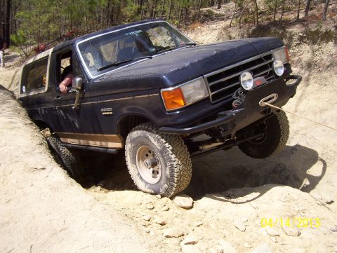 1990 Ford Bronco - A dark blue Ford Bronco navigates a challenging off-road obstacle, its front tire deep in a sandy trench.