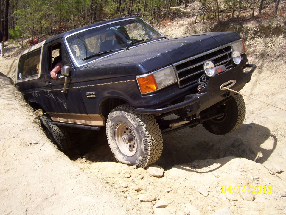 Dark blue Ford Bronco with a tan stripe, stuck in a dirt ditch, with a winch cable attached to the front bumper