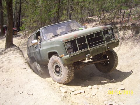 1990 Ford Bronco - Olive drab off-road SUV navigates a steep dirt trail with a second vehicle in the background.