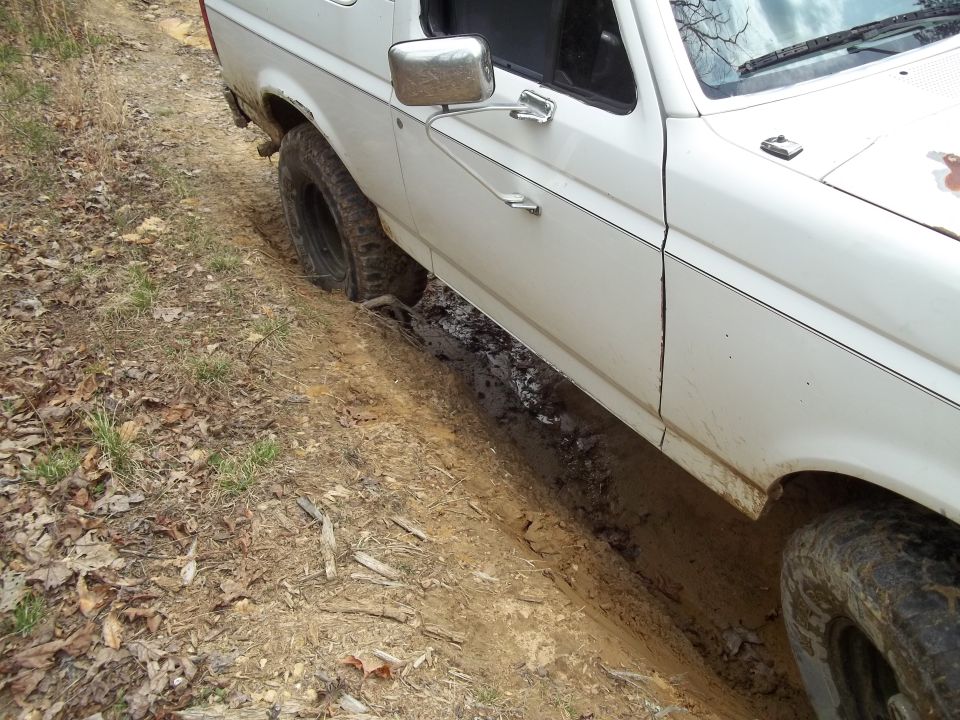 White SUV with one wheel in a muddy rut on a dirt path