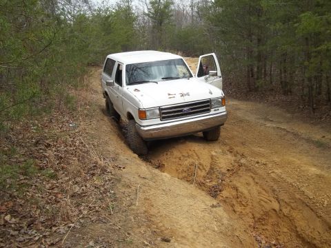 1990 Ford Bronco - A white Ford Bronco navigates a challenging off-road trail with its driver's door ajar.