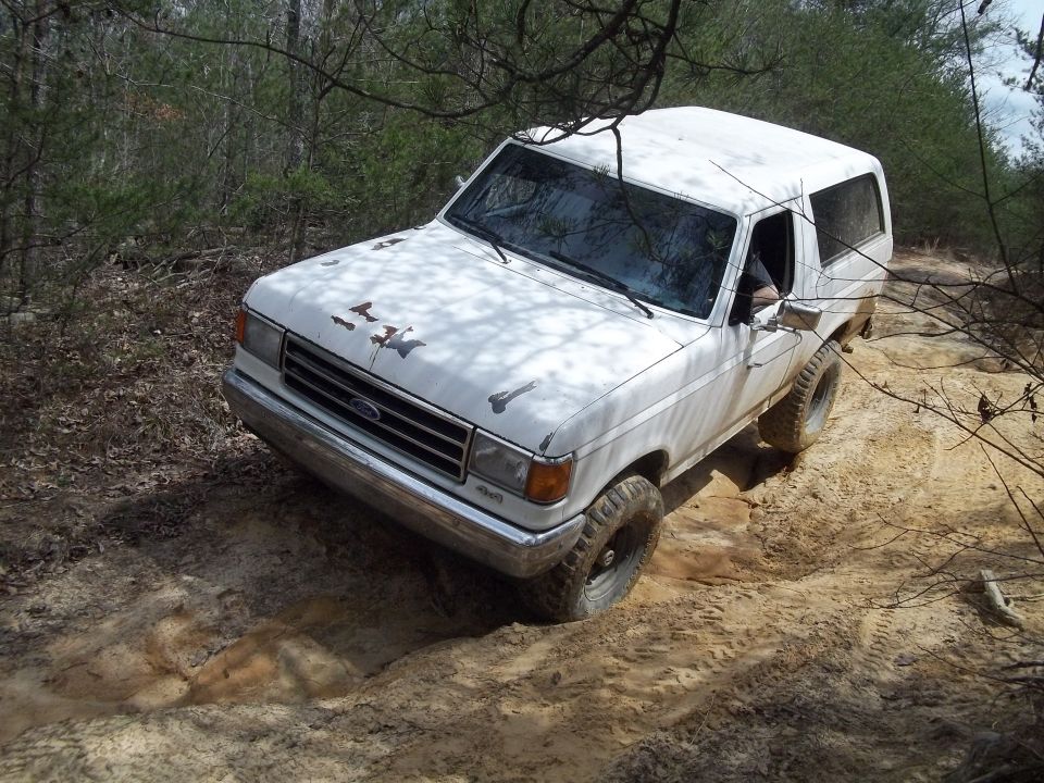 White 1987 Ford Bronco on a dirt trail, front-left quarter view, with rust spots on hood