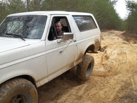 1990 Ford Bronco - White 1990 Ford Bronco navigates a muddy off-road path with trees in the background.