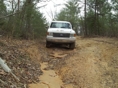 1990 Ford Bronco - A white Ford truck navigates a muddy, rutted path in a wooded area.